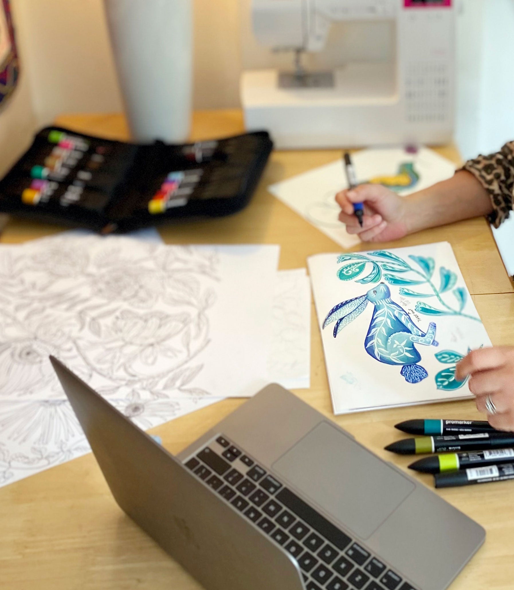 Person coloring a hare design on paper with markers next to a laptop on a wooden table.